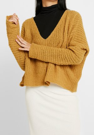 Femme ajustant la manche d'un pull épais ample jaune moutarde porté sur un col roulé noir, associé à une jupe blanche côtelée.