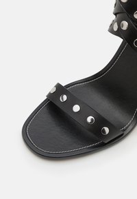 Μαύρο δερμάτινο σανδάλι με φαρδιές λωρίδες και ασημένιους μεταλλικούς στρόγγυλους συνδέσμους, τετράγωνη μύτη και αντίθετη λευκή ραφή στη σόλα.