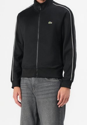 Mann trägt eine schwarze Zip-up-Jacke mit weißen Streifen an den Ärmeln und einem kleinen Krokodillogo, kombiniert mit grauen Jeans.