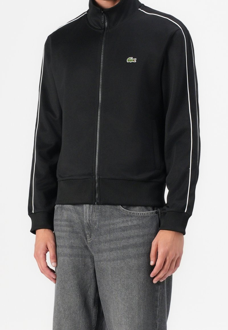 Homme portant une veste noire zippée avec des bandes blanches sur les manches et un petit logo crocodile, associée à un jean gris.