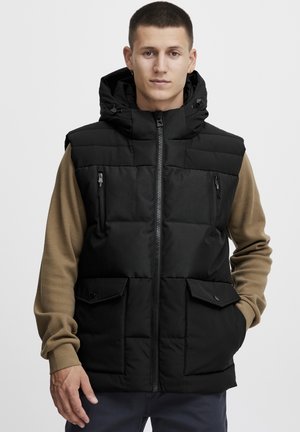 Black padded vest with a zip front, side pockets, and a hood. Features a contrast brown long-sleeve shirt underneath. Smooth texture.