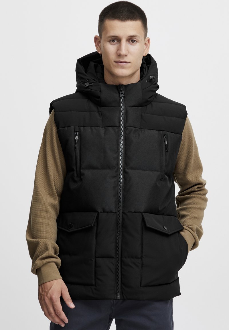 Gilet imbottito nero con zip frontale, tasche laterali e cappuccio. Sotto presenta una maglietta a maniche lunghe marrone in contrasto. Tessuto liscio.