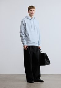 Φωτεινή γαλάζια φούτερ με κουκούλα και μπροστινή τσέπη, συνδυασμένη με μαύρο παντελόνι φαρδιάς γραμμής. Το μοντέλο κρατάει μια μαύρη τσάντα. Απλός σχεδιασμός με ελάχιστη σήμανση.