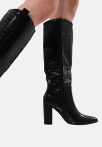 Botas negras hasta la rodilla, hechas de un material brillante con un estampado de cocodrilo. Presentan una punta afilada y un tacón ancho, con un cierre de cremallera en el lateral.
