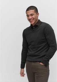 Mörkgrå v-ringad tröja med långa ärmar, bärs över en kragtröja i brunt och parats med ljusbruna byxor. Slät yta, minimal design.