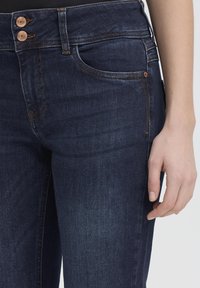 Primer plano de la cintura y la parte superior del muslo de unos vaqueros de mezclilla azul oscuro con la mano apoyada junto a la pierna, con botones de cobre y costuras naranjas.