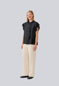 Blusa preta de volantes com mangas curtas, frente abotoada; acompanhada por calças largas texturizadas em creme e sapatos pretos, fundo sólido.