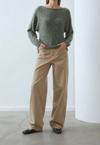 Grön stickad tröja med lös passform, med små detaljer. Beigefärgade vida byxor i kraftigt tyg, kombinerade med mörka platta skor.