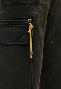 Μαύρο παντελόνι cargo από ύφασμα με φερμουάρ και κίτρινο λουράκι με ξύλινο χάντρα. Απαλή υφή και κομψή εφαρμογή.