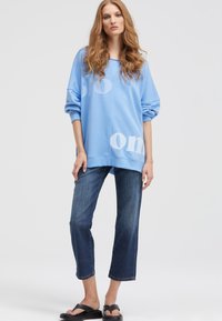 Oversized light blue sweatshirt with white text, paired with dark denim cropped jeans. Model wears black sandals. Smooth texture, relaxed fit.