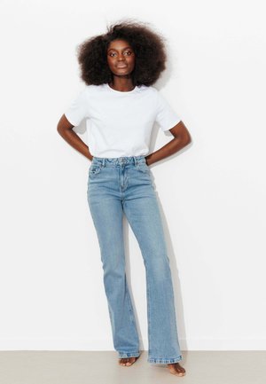 Femme confiante aux cheveux afro, debout pieds nus contre un mur blanc, portant un t-shirt blanc uni et un jean taille haute bleu clair.
