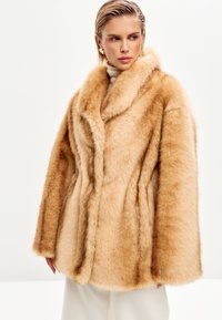 Lysebrun kunstpelsfrakke med bred krave, der har en blød tekstur, afslappet pasform og diskrete sømdetaljer. Bæres over en lys turtleneck.