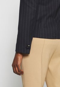 Ręka o ciemnej karnacji spoczywająca na boku, ubrana w granatową marynarkę w prążki i beżowe spodnie dopasowane, na tle jasnego, gładkiego tła.