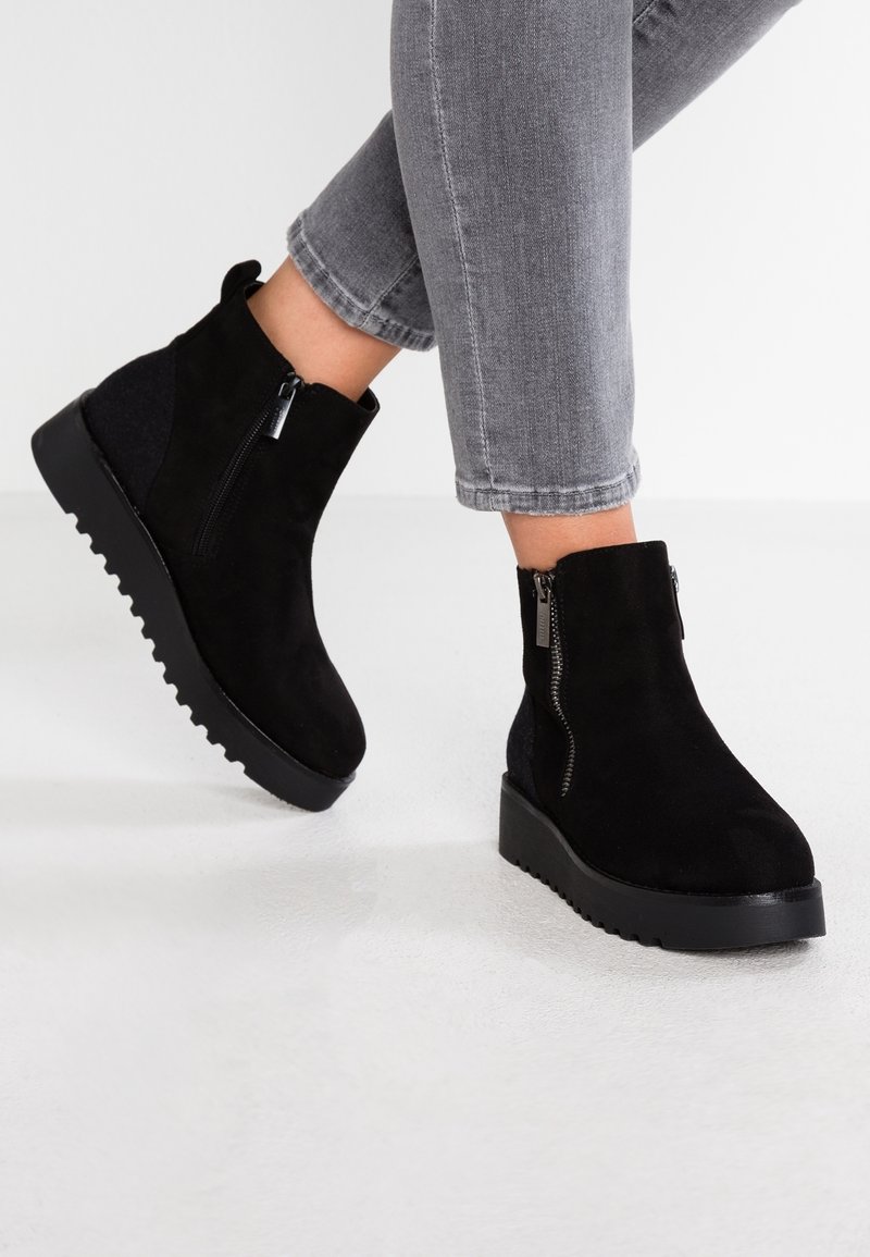 Personne portant des bottines en daim noir avec des fermetures éclair latérales et des semelles rainurées, associées à un jean gris court sur un sol blanc.