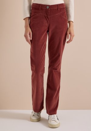 Femme portant un pantalon en velours côtelé couleur rouille, coupe droite, avec poches, et des baskets blanches sur un fond beige uni.
