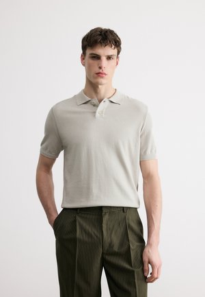 Ung mand iført en lys beige polo og mørkegrønne bukser med nålestriber, stående med den ene hånd i lommen mod en ensfarvet baggrund.