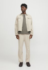 Beige denimjakke med to brystlommer, olivengrøn T-shirt under, cremede bukser og brune snørebåndssko. Minimalistisk design og afslappet pasform.