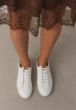 Ben iført en brun blondernederdel og hvide sneakers med leopardmønsterdetaljer på en lys grå gulv.