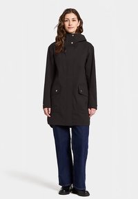 Zwarte waterdichte jas met capuchon, twee voorzakken en drukknopdetails. Gedragen over donkerblauwe wijde jeans en zwarte schoenen.