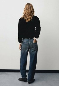 Pull texturé noir, jean bleu taille haute à coupe ample, et bottines noires. Vue de dos, cheveux ondulés descendant au-delà des épaules.