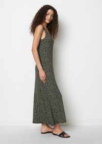Maxikleid in Dunkelgrün mit einem weißen Muster. Es hat dünne Träger und eine fließende Silhouette, kombiniert mit schwarzen Rutschsandalen.