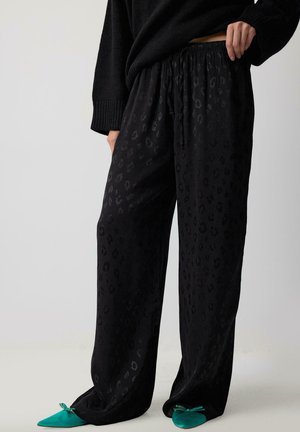 Persoon die zwarte, loszittende broek met patroon en elastische taille draagt, met groene puntige platte schoenen, staand tegen een effen lichte achtergrond.