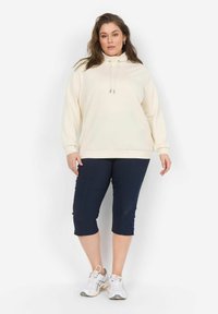 Sudadera de manga larga color crema claro con cuello alto y cordón. Combinada con pantalones capri azul marino. Zapatos blancos con detalles en gris.