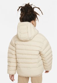 Chaqueta acolchada beige con capucha, con rayas horizontales acolchadas y un ajuste ligeramente holgado. Textura suave y dobladillo redondeado.