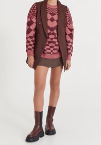 Brown tailored vest over a pink patterned sweater, paired with a pleated brown mini skirt and brown ankle boots with thick soles.