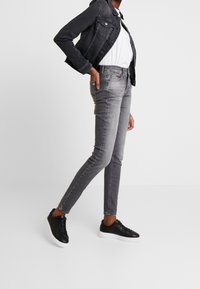 Jeans ajustados gris con un acabado desgastado, combinados con una camisa blanca y una chaqueta de mezclilla negra, acentuados por zapatillas negras.