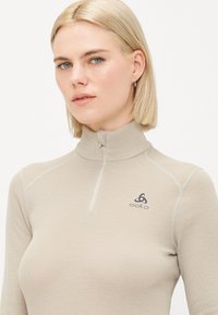 Beige long-sleeve top with a high collar, featuring a half-zip and grey stitching. Includes the odlo logo on the left side.