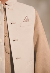 Veste couleur crème avec une finition texturée, dotée d'une poche contenant un mouchoir à motif et de boutons en bois contrastant sur une chemise en-dessous.