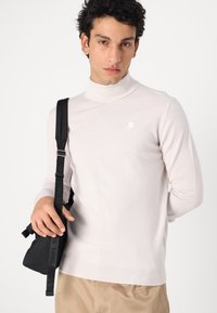 Hellgrauer Rollkragenpullover aus weichem Stoff mit kleinem Logo auf der Brust. Das Modell hält eine schwarze Umhängetasche mit Texthinweisen.