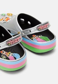 Crocs argentés avec un intérieur noir présentant le design des Powerpuff Girls. La semelle multicolore comprend des rayures vertes, roses et bleues.