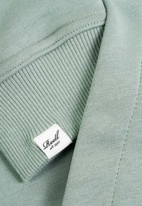 Light green ribbed sweater cuff with a white fabric label reading "Reell est. 1997" sewn near the seam.
