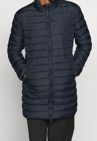 Marinblå pufferjacka med fullt framdraget dragkedja, horisontellt quiltat mönster, hög krage och sidofickor. Slät, lättviktsmaterial.