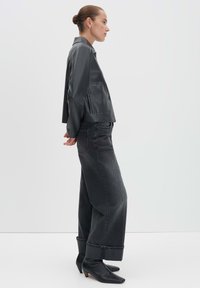 Graue satinierte Jacke mit Kragen, kombiniert mit weit geschnittenen schwarzen Jeans aus Denim mit Umschlägen. Das Modell trägt spitze schwarze Stiefeletten.