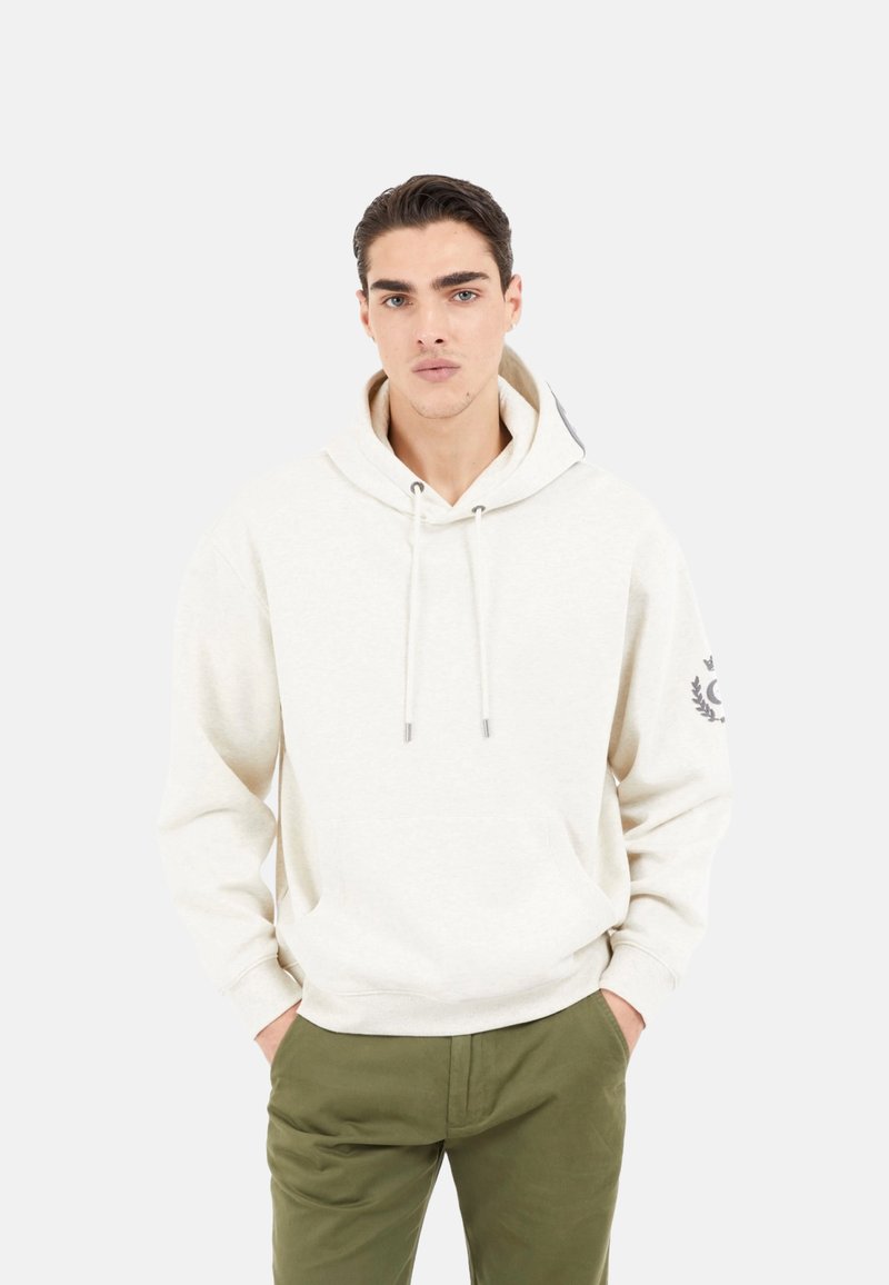 Sudadera de color beige claro con un bolsillo tipo canguro, cordones plateados y un gráfico en la manga izquierda. Combinada con pantalones de color verde oliva.