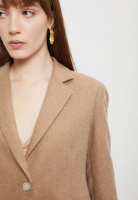 Bež vuna sako s reverima i jednim gumbom, koji prikazuje glatku teksturu. Zlatne naušnice s privjeskom s složenim diskovima.