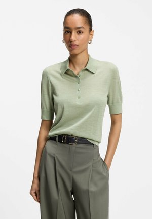 Femme portant un polo vert clair à manches courtes rentré dans un pantalon taille haute couleur olive avec une ceinture noire.