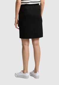 Femme portant une jupe noire jusqu'au genou, un haut sans manches à rayures et des baskets blanches, se tenant de dos par rapport à la caméra.