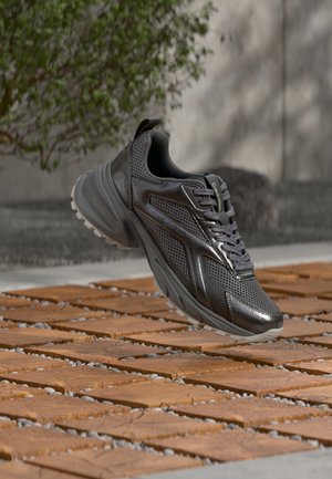 Chaussure de sport noire avec un design texturé et une semelle épaisse flottant au-dessus d'une surface extérieure en bois et pierre à motifs, avec un arbre et un mur en arrière-plan.
