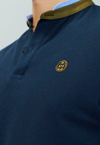 Mies, joka käyttää tummansinistä polo-paitaa, jossa on ruskea kauluskaitale ja pieni pyöreä kultainen emblem vasemmalla rinnalla.