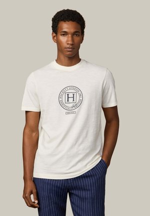 Jonge man draagt een wit T-shirt met het logo "Hackett London 1983 Essentially British" en marineblauwe broek met krijtstrepen, poserend tegen een neutrale achtergrond.
