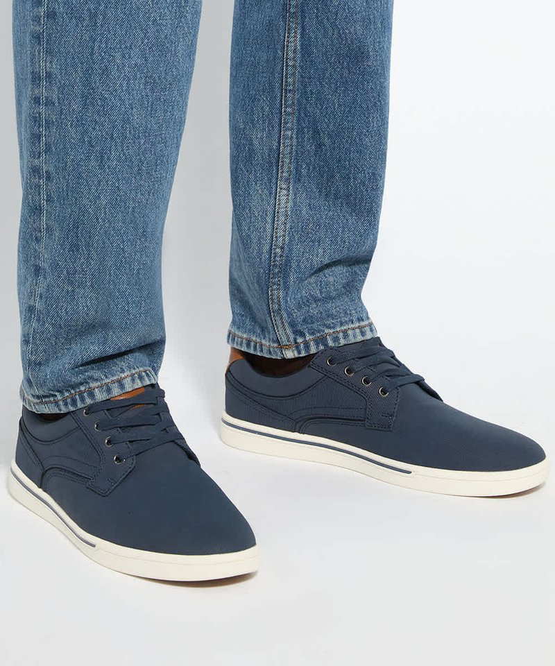 Personne portant un jean bleu et des chaussures décontractées lacées bleu marine avec des semelles blanches, debout sur un fond clair.