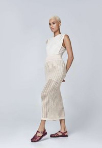 Kremowy top bez rękawów zestawiony z dzianinową spódnicą w beżowym kolorze z otwartym wzorem, dopełniony bordowymi sandałami z paskowym wzorem.