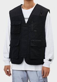 Chaleco utilitario negro de tela con múltiples bolsillos, detalles de malla y cremalleras. Presenta un diseño sin mangas y una construcción en capas.