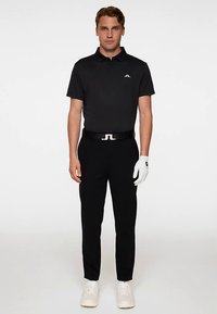 Svart pikétröja med korta ärmar, matchande svarta byxor, vitt bälte med logotyp, vit golfhandske och vita sneakers. Enfärgad bakgrund.