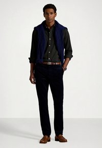 Granatowa kamizelka w warkoczowy splot noszona na ciemnej koszuli na guziki, zestawiona z ciemnymi spodniami i brązowymi skórzanymi butami. W stylizacji znajduje się także brązowy pasek i zegarek.