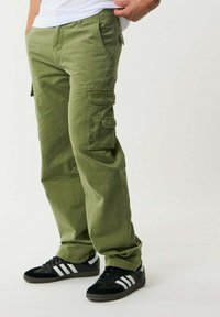 Pantaloni cargo verde oliva con tasche laterali, vestibilità comoda e logo visibile sulla gamba. Abbinati a scarpe sportive nere con strisce bianche.